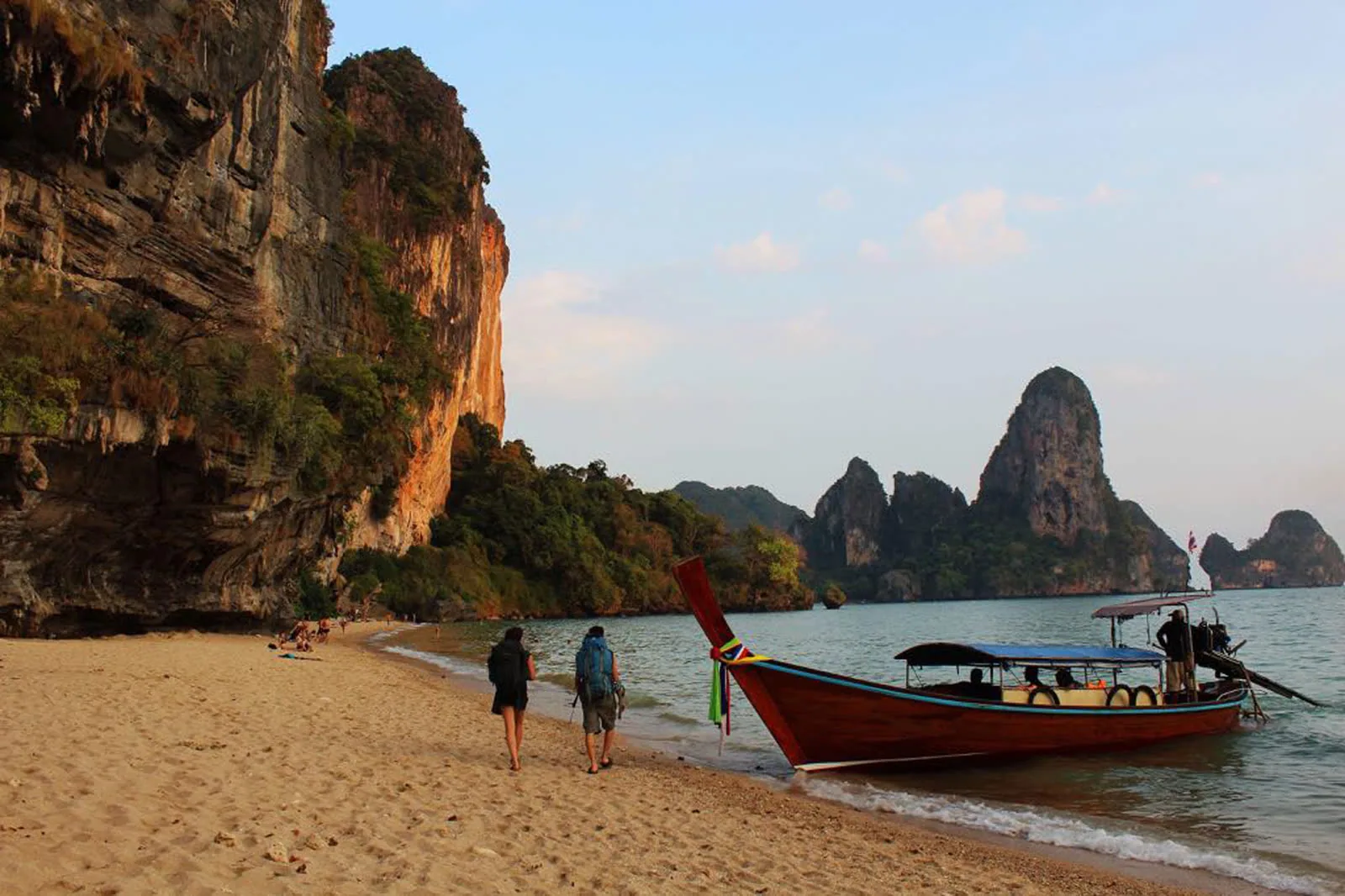 Two travelers walk beside a long-tail boat on a Krabi beach below tall limestone cliffs.